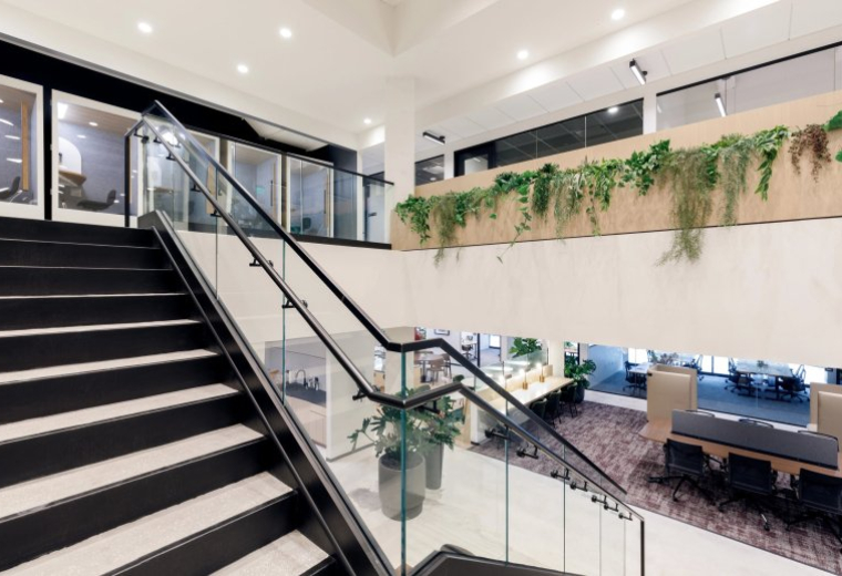 Spacious bi-level lobby at 3390 Mary Street with a black staircase, glass railings, and lush hanging greenery.