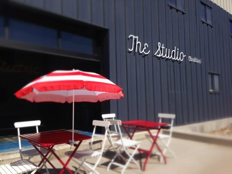 Exterior entrance of the dark metal-clad building at 3550 Frontier Avenue A2 with red patio umbrellas.