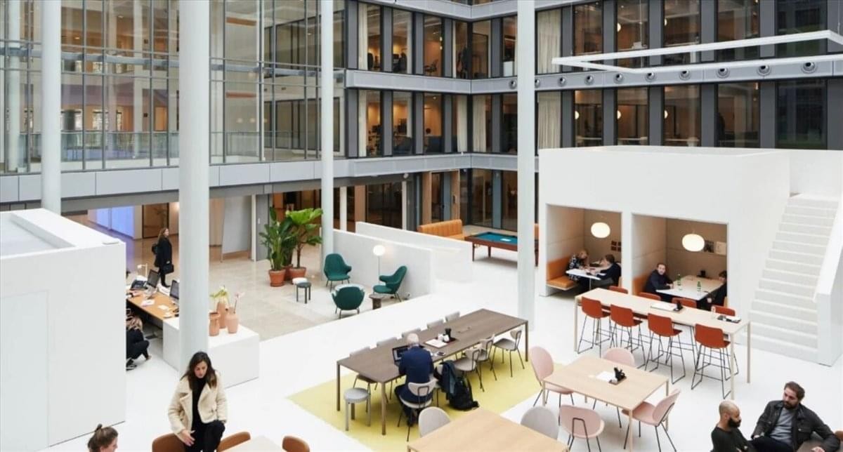 Bright multi-level communal atrium with various seating areas, tables, and indoor greenery.