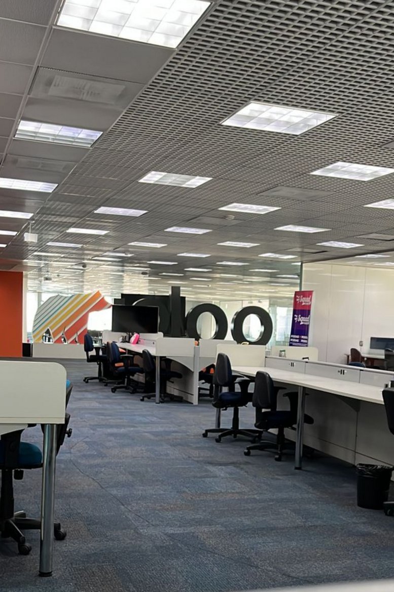 Large open-plan office space with rows of white desks, blue carpet, and a modern grid ceiling.