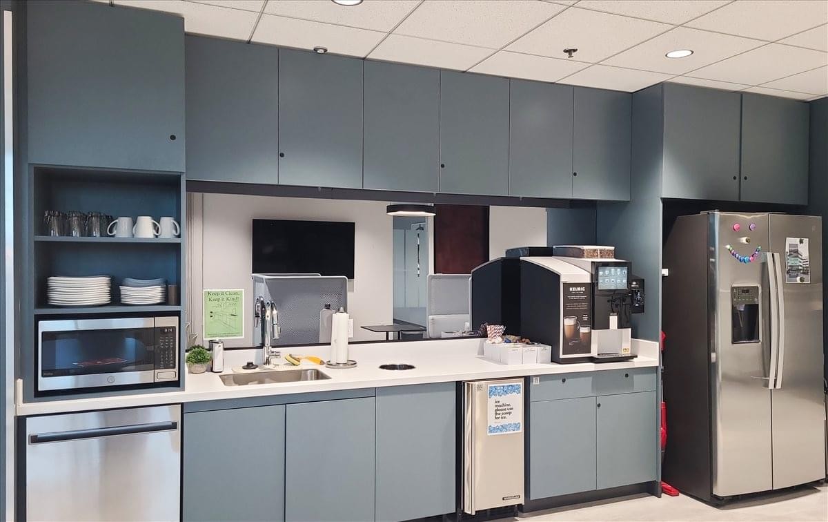 Modern communal kitchen and break area with blue cabinetry, coffee machine, and full-sized refrigerator.