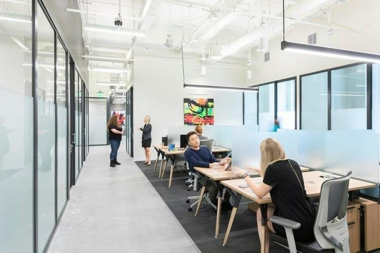 Open-plan office with desks and glass-walled private suites along the corridor.