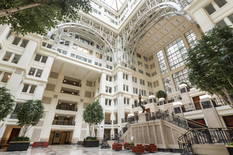 Grand atrium lobby of 601 13th Street NW with a multi-story glass ceiling, white arches, and indoor trees.