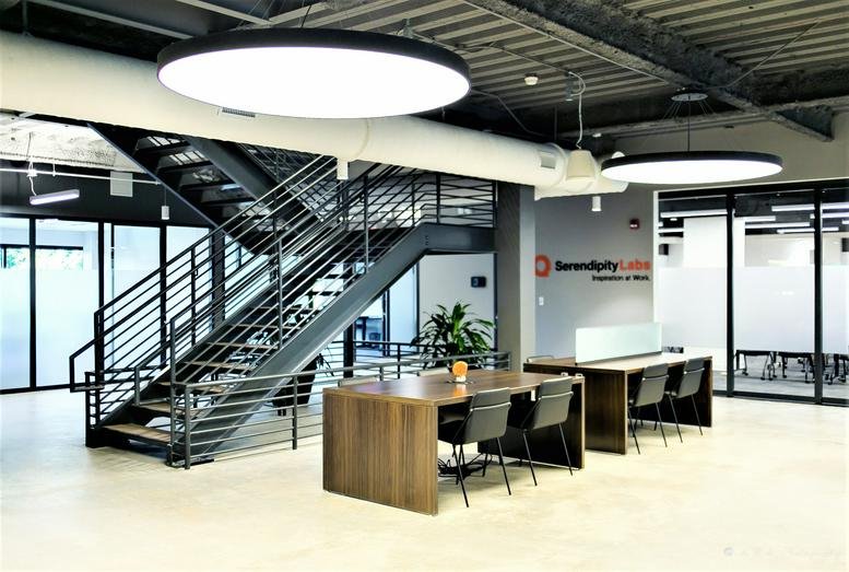 Spacious coworking area with wooden tables, black chairs, and a metal staircase under large circular lights.