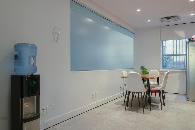 Dining area with a water cooler and a table surrounded by white chairs against a minimalist wall.