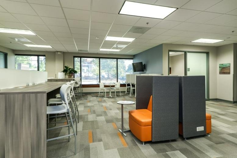Spacious communal lounge with orange privacy pods, a long wooden high-top desk, and tall windows.