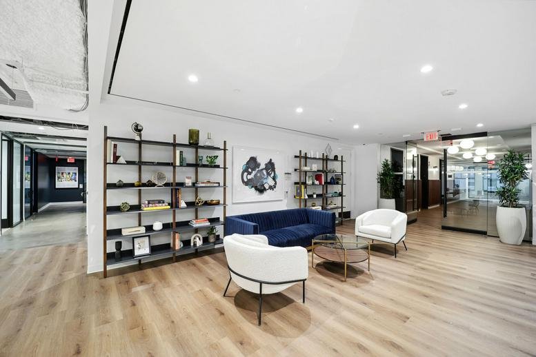 Bright communal lounge area featuring a blue velvet sofa, white armchairs, and wooden floors.