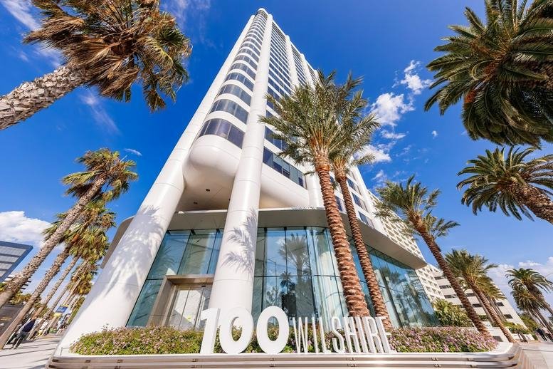 Ground-up view of the 100 Wilshire Blvd white skyscraper facade with palm trees and a blue sky.