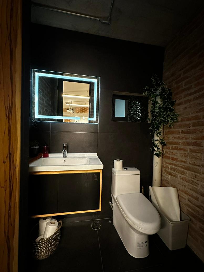 Sleek restroom with a black wall, a large backlit mirror, and exposed brick accents.