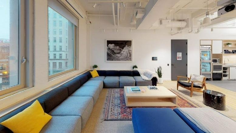 Spacious communal lounge with a grey sectional sofa, wooden floors, and city views.