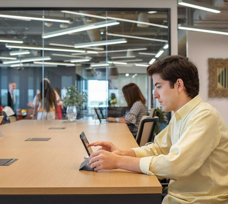 Professional working at a wooden communal table with a view of a busy office through glass partitions.