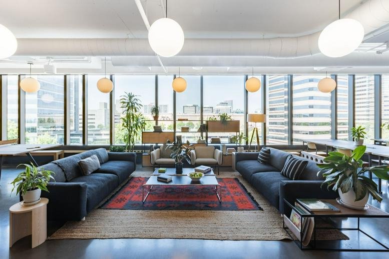 Spacious lounge with blue sofas, colorful rugs, indoor plants, and floor-to-ceiling windows.
