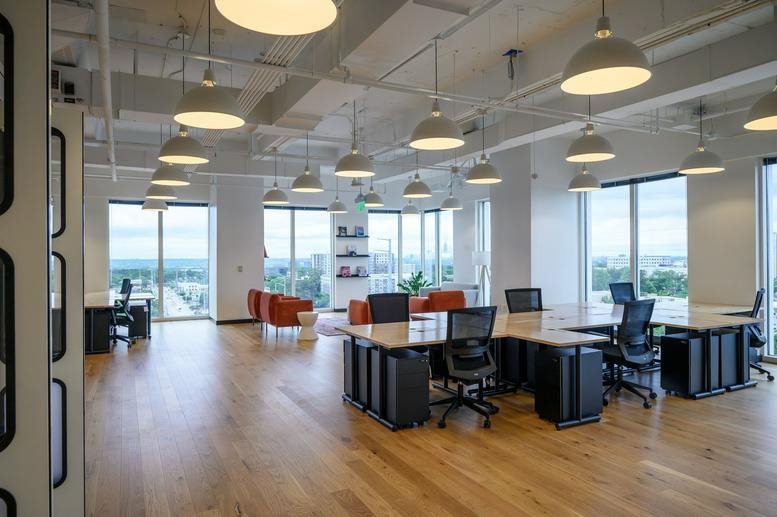 Spacious open-plan workspace with multiple desk clusters, ergonomic chairs, and city views.
