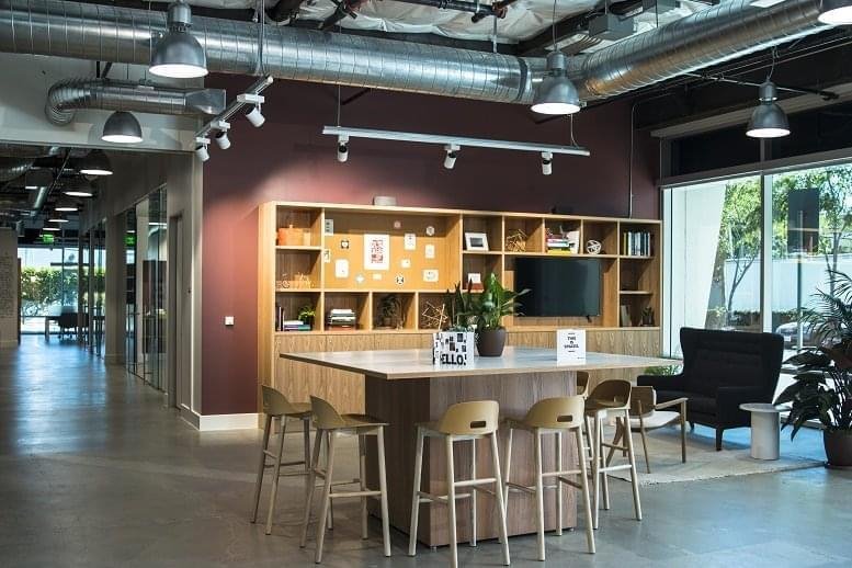 Spacious communal lounge featuring a wooden island table, bookshelf wall, and industrial ceilings.