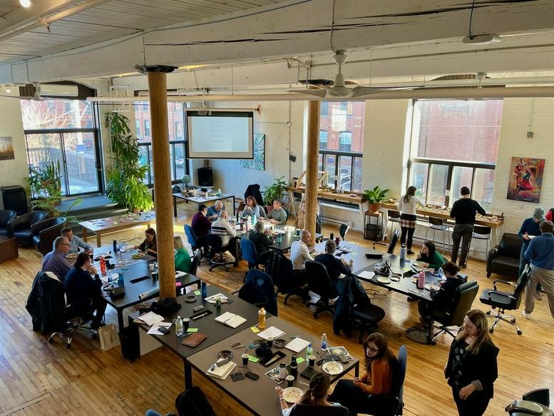 Large shared office space filled with people working at long communal desks under a high ceiling.