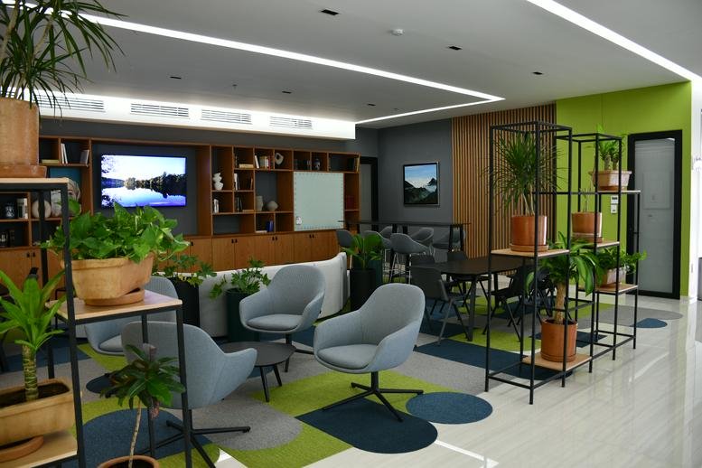 Spacious communal lounge at Dario Business Center with grey armchairs, indoor plants, and a wall-mounted TV.