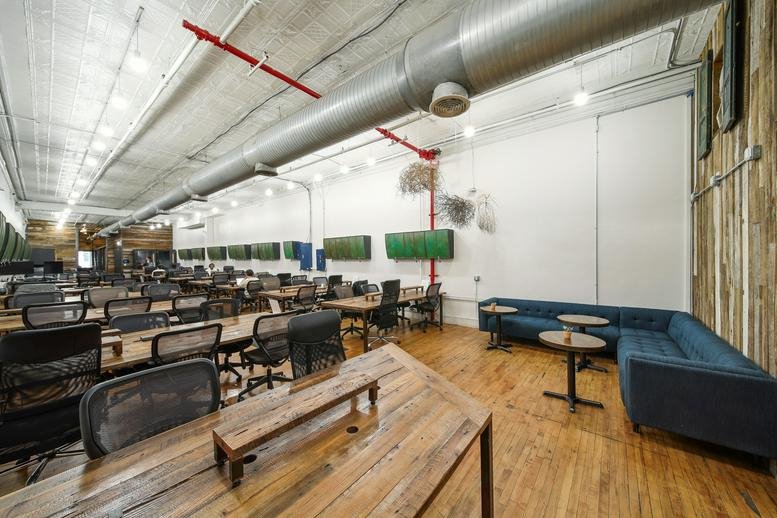 Wide-angle view of the open-plan office showing desks, a blue sofa, and industrial ceiling ducts.