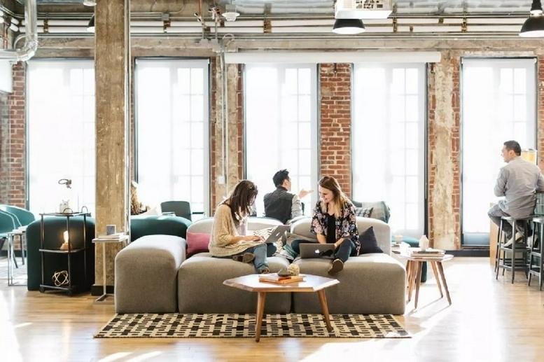 Spacious communal lounge with exposed brick, large windows, and people working on comfortable sofas.