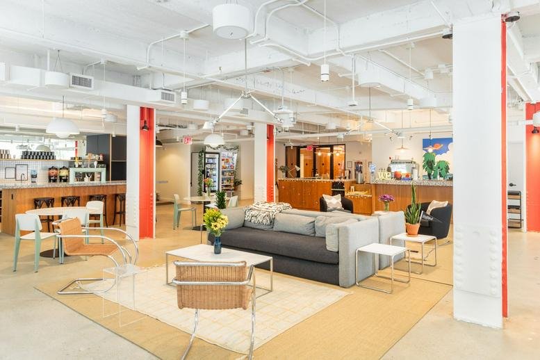 Bright, spacious coworking lounge with grey sofas, white tables, and orange structural columns.