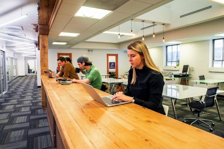 Spacious coworking area with a long wooden high-top desk and bright overhead lighting.