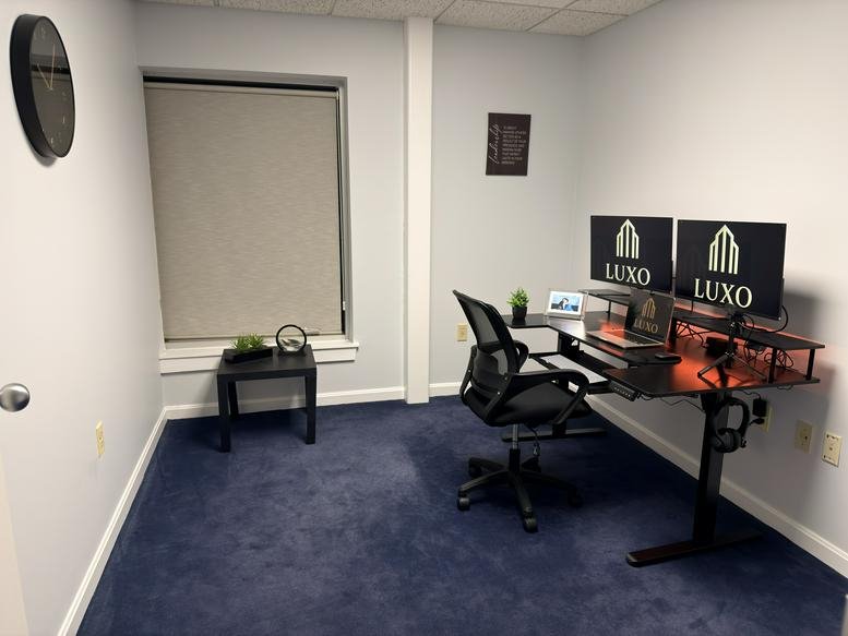 Private office featuring a black sit-stand desk, mesh chair, and dark blue carpeting.