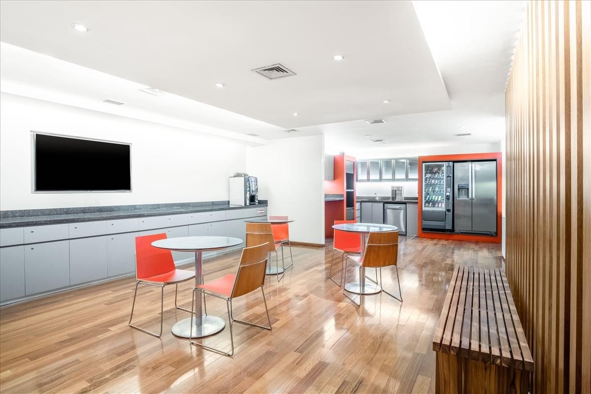 Communal breakroom with cafe tables, red chairs, wooden flooring, and a refreshment area.