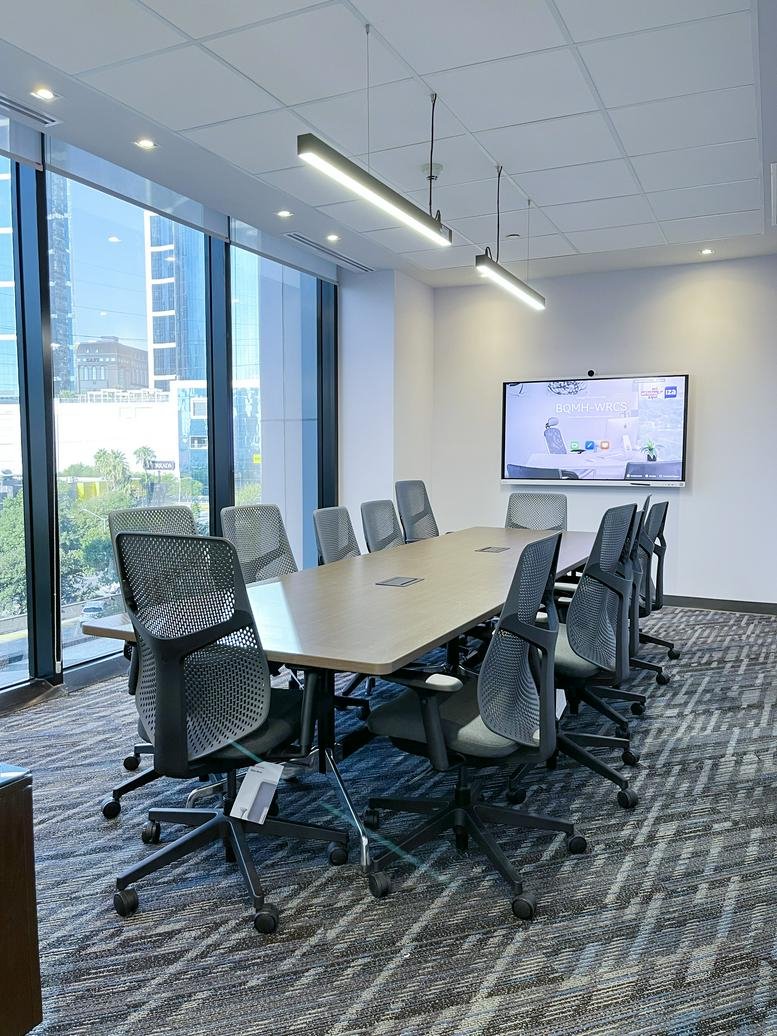 Sunlit meeting space with a large wooden table, yellow chairs, and city views through floor-to-ceiling glass.