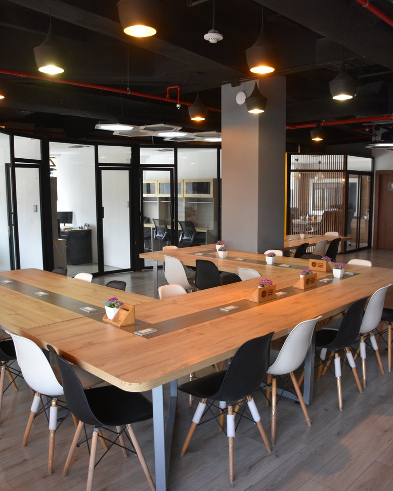 Spacious communal coworking area at Edificio 100A with warm wood U-shaped desks and modern pendant lighting.