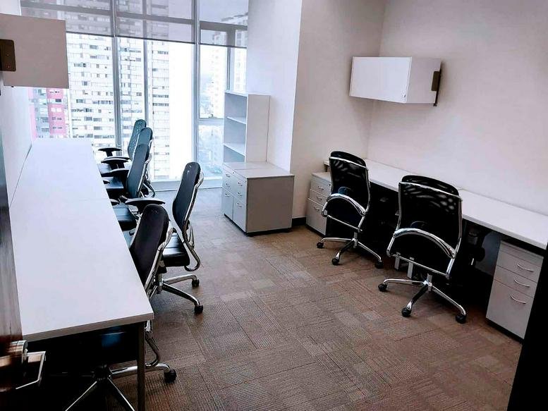 Shared office workspace with white desks and black chairs positioned near bright windows.