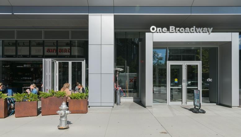 Ground level exterior entrance and facade of One Broadway with outdoor seating and plants.