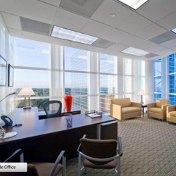 Sun-drenched executive corner office with floor-to-ceiling windows, an L-shaped desk, and lounge seating.