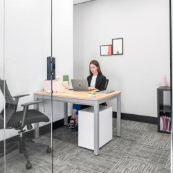 Modern glass-partitioned private office with a desk, shelving, and a person working.