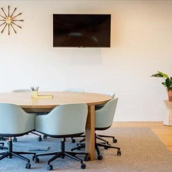 Minimalist meeting room featuring a round wooden table, light blue chairs, and a wall-mounted screen.