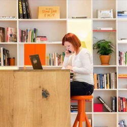 Quiet workspace with a person at a high desk in front of a large white shelving unit full of books.