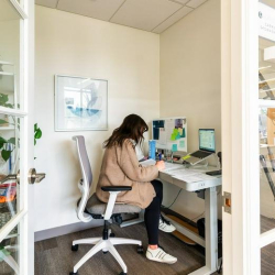Private glass-walled office with a standing desk and ergonomic chair.